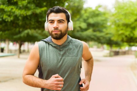 Foto de Active handsome man listening to music with headphones while running and exercising at the park - Imagen libre de derechos