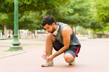 Téléchargez les photos : Hispanic young man tying his shoes and preparing to go for a run in the park with headphones - en image libre de droit
