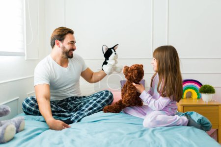 Téléchargez les photos : Cheerful smiling father in pajamas playing puppets with her cute daughter after waking up in bed - en image libre de droit