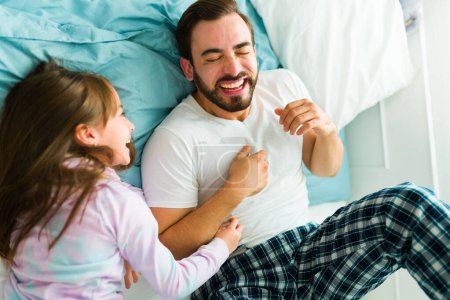 Téléchargez les photos : Cute little girl tickling her dad's stomach and laughing together having fun in pajamas after waking up - en image libre de droit