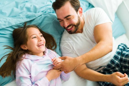 Foto de Loving happy father tickling her daughter and laughing together after waking up in bed during the morning - Imagen libre de derechos