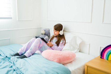 Photo for Adorable little girl in pajamas relaxing in bed with headphones using the tablet to watch online videos - Royalty Free Image