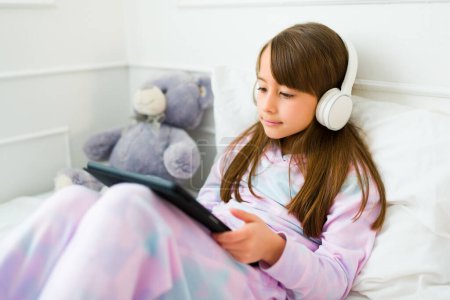 Photo for Cute little girl with headphones watching videos on the tablet or playing online videogames in bed wearing pjs - Royalty Free Image