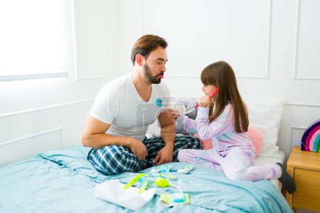 Photo for Beautiful child using a toy stethoscope and playing doctor with her sick father in pajamas - Royalty Free Image
