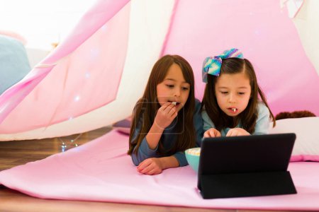 Téléchargez les photos : Caucasian children relaxing inside a pink teepee eating popcorn watching a movie on the tablet together - en image libre de droit