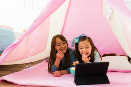 Photo for Happy little girls laughing having fun while watching a children's movie on the tablet and eating popcorn - Royalty Free Image