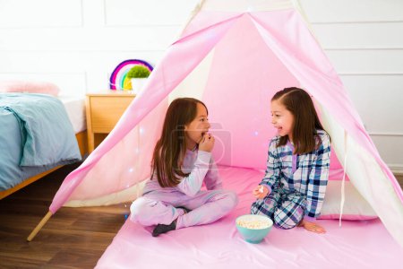 Photo for Happy little girls in pajamas playing during a fun sleepover eating popcorn while talking in a pink teepee - Royalty Free Image