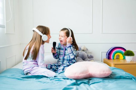 Foto de Happy little girls looking excited while playing karaoke and singing wearing headphones during a sleepover - Imagen libre de derechos