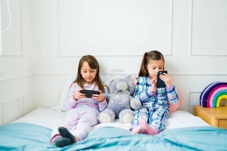 Photo for Children friends in pajamas resting in bed during a slumber party while playing video games on the smartphone - Royalty Free Image