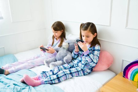 Foto de Little girls friends wearing pjs in bed having a slumber party playing on the smartphone - Imagen libre de derechos