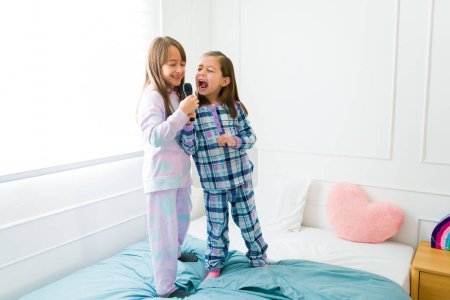 Foto de Fun children and best friends in pajamas playing karaoke singing standing in bed during a slumber party - Imagen libre de derechos