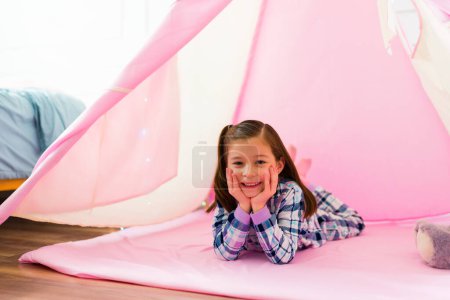 Téléchargez les photos : Adorable little girl relaxing looking happy in pajamas while playing inside a pink teepee in her bedroom - en image libre de droit