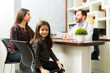 Photo for Cute hispanic young kid smiling with her mother talking with the pediatrician at the doctor's office after  medical check up - Royalty Free Image
