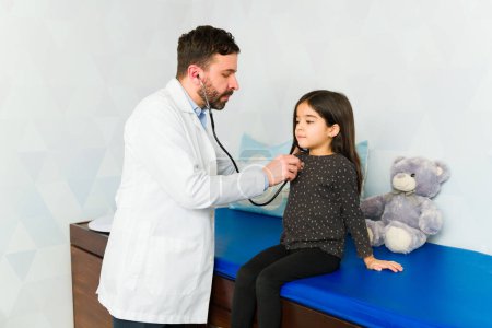 Foto de Médico pediatra caucásico usando un estetoscopio mientras revisa y hace un examen médico en un niño pequeño latino - Imagen libre de derechos