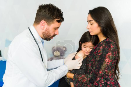Foto de Madre hispana sosteniendo a su asustada hija enferma recibiendo una vacuna contra la medicina en el consultorio del médico - Imagen libre de derechos