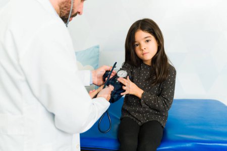 Téléchargez les photos : Triste petite fille regardant la caméra lors d'un examen médical à l'aide d'un manomètre chez le médecin - en image libre de droit