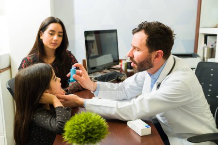 Photo for Pediatrician explaining how to use an asthma inhaler to a little kid with her mother sitting at his desk office - Royalty Free Image