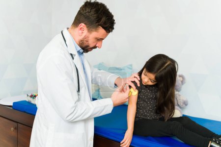 Foto de Caucasian pediatrician doctor putting an injection vaccine or sensory device to a sick young child - Imagen libre de derechos