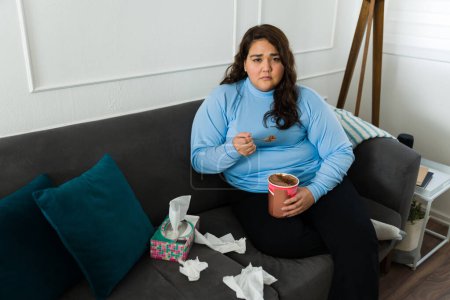 Téléchargez les photos : Coeur brisé femme obèse regardant déprimé et triste tout en mangeant de la crème glacée au chocolat sur le canapé - en image libre de droit