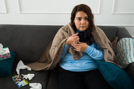 Foto de Mujer deprimida con sobrepeso haciendo contacto visual mientras bebe té caliente llorando mirando triste y solo en la sala de estar - Imagen libre de derechos