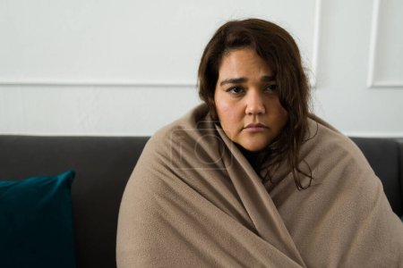 Photo for Hispanic overweight woman feeling sad and lonely wrapped in a blanket while suffering alone from depression and mental health problems - Royalty Free Image