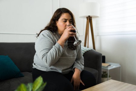 Photo for Latin fat woman drinking wine and looking tired and sad while feeling depressed and lonely at home - Royalty Free Image