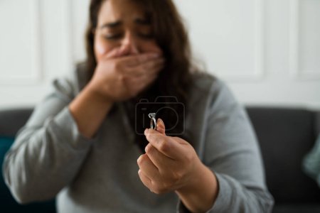 Téléchargez les photos : Triste jeune femme pleurant en regardant sa bague de fiançailles tout en se sentant déprimée par sa rupture brisée avec son ex-fiancé - en image libre de droit