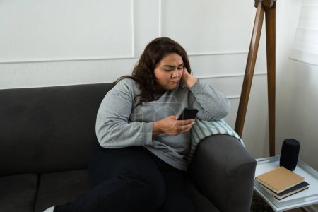 Téléchargez les photos : Gros latin femme à la recherche triste et se sentir comme pleurer tout en regardant les médias sociaux ou des photos sur son téléphone de son ex-petit ami - en image libre de droit