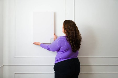 Photo for Attractive fat woman seen from behind putting a picture frame or mockup on the wall while decorating her home - Royalty Free Image
