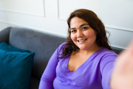 Téléchargez les photos : Point de vue personnel d'une femme hispanique grosse prenant un selfie et souriant tout en se reposant sur le canapé à la maison - en image libre de droit