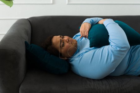 Foto de Sad obese woman hugging a pillow while lying on the sofa and crying missing her boyfriend after a breakup feeling lonely - Imagen libre de derechos
