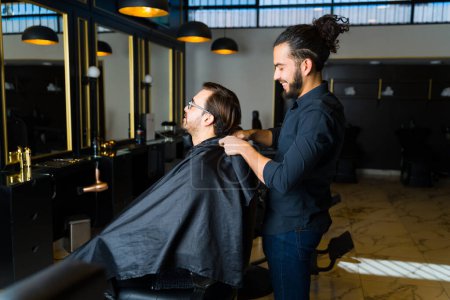 Téléchargez les photos : Joyeux barbier souriant tout en travaillant avec un client caucasien obtenir une nouvelle coupe de cheveux au salon de coiffure - en image libre de droit