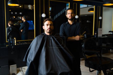 Téléchargez les photos : Attractive young man with a male hairstylist at the barber shop ready to trim his beard and get a haircut - en image libre de droit