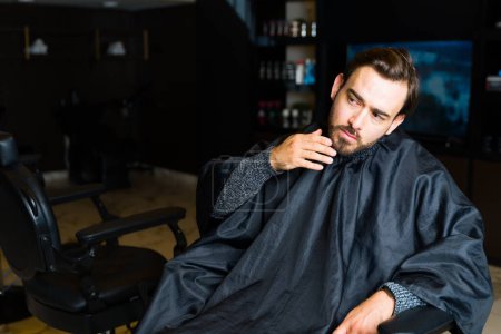 Téléchargez les photos : Jeune homme caucasien au salon de coiffure vérifiant sa barbe et ses cheveux en attendant d'obtenir un service de toilettage - en image libre de droit