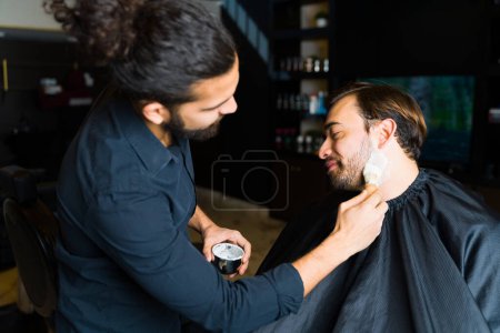 Photo for Hispanic barber putting shaving cream on a happy man shaving his beard and cutting his hair at the barber shop - Royalty Free Image