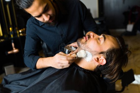 Foto de Male barber putting shaving cream on the face of a caucasian customer shaving his beard and getting a grooming service - Imagen libre de derechos