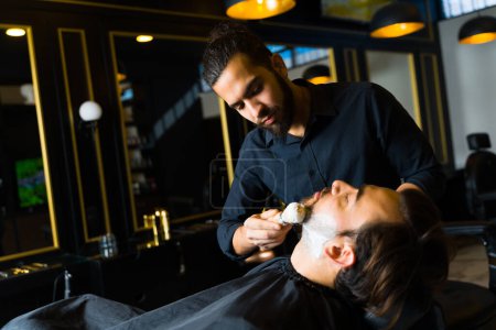 Photo for Attractive barber putting shaving cream and grooming the beard of a male customer at the barber shop - Royalty Free Image