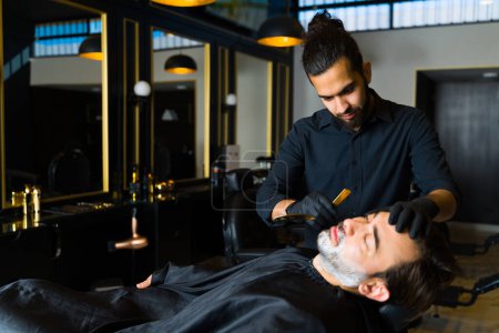 Foto de Handsome hispanic barber using cream to shave the beard of a man customer at a classy barber shop - Imagen libre de derechos