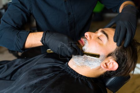Photo for Close up of a barber using a razor blade to shave the beard of a relaxed caucasian man at the salon - Royalty Free Image