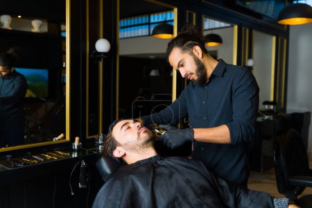 Téléchargez les photos : Beau barbier professionnel taillant et détaillant la barbe d'un client masculin attrayant dans un salon de coiffure chic - en image libre de droit