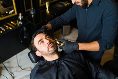 Foto de High angle of young man and customer trimming his beard and getting grooming services with a professional barber - Imagen libre de derechos