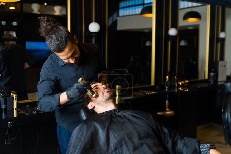 Photo for Male barber shaving the beard of a caucasian client while working at a sophisticated barber shop - Royalty Free Image