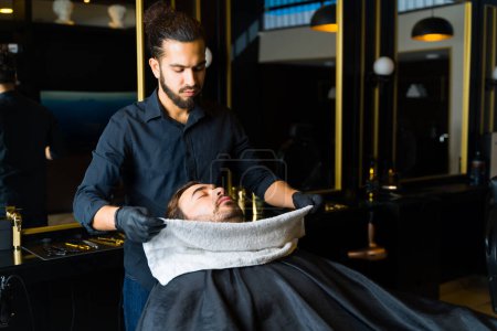 Photo for Professional latin barber putting a hot towel on a customer to open his pores before shaving his beard - Royalty Free Image
