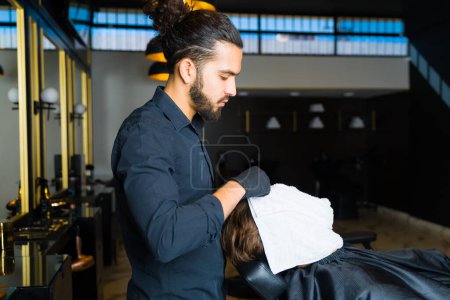 Foto de Professional hispanic barber putting a hot towel on a relaxed young man to help the skin before shaving his beard - Imagen libre de derechos