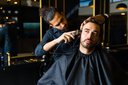 Foto de Professional barber using a machine while cutting the hair of a caucasian male client at the hair salon - Imagen libre de derechos
