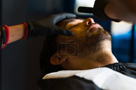Téléchargez les photos : Attrayant caucasien homme relaxant tout en rasant sa barbe avec une lame de rasoir par un coiffeur professionnel - en image libre de droit