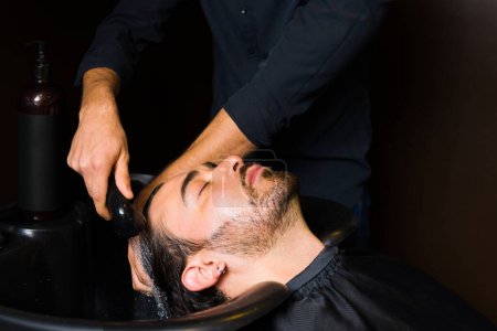 Foto de Young relaxed man getting his hair washed at the hair salon before getting a new trendy haircut from the barber - Imagen libre de derechos