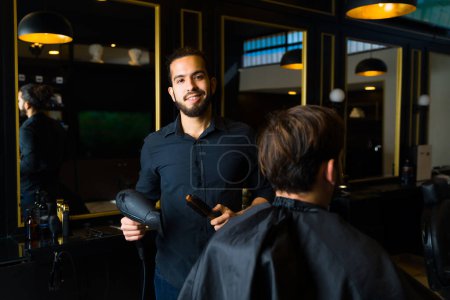 Photo for Cheerful attractive man smiling working as a hairstylist using a blow dryer while giving a haircut to a client - Royalty Free Image