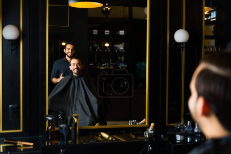 Foto de Cheerful male hairstylist and young man smiling looking in the mirror after getting a new trendy haircut at the salon - Imagen libre de derechos