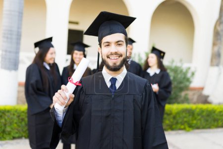 Téléchargez les photos : Jeune homme hispanique attrayant montrant son diplôme d'études supérieures après avoir assisté à sa cérémonie de remise des diplômes sur le campus du collège - en image libre de droit
